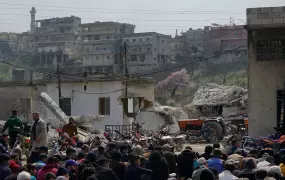 Location: Maland village, Idlib. A group of Syrian Muslims gather to perform Friday prayers. All people are seated and in the backdrop we see building from the village.